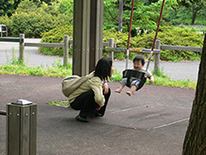 写真:幼児用のブランコに乗りご機嫌のあかちゃん。地面にしゃがんであかちゃんと笑顔を交わすお母さん