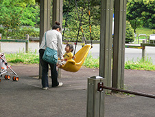 写真:背もたれ付きブランコに乗って笑顔になった小さな女の子。お母さんが正面に立ちブランコをそっと押す