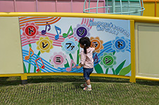 写真:壁に取り付けられた幅180センチほどのパネルとその前に立つ女の子。パネルに描かれた8つの花にはそれぞれドレミファソラシドの文字と、その上に「花のうえに手をのせると音が出るよ!」の説明