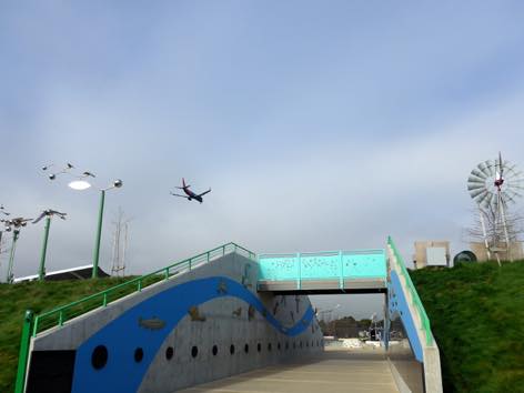 写真:土手の間を貫くアプローチ。上にはブリッジが架かり、空に飛行機の姿。