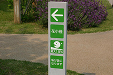 写真:園路の分かれ道にある道標。緑のパネルに白抜きの字。大きな矢印や勾配が急になることを示すピクトグラムも示されている